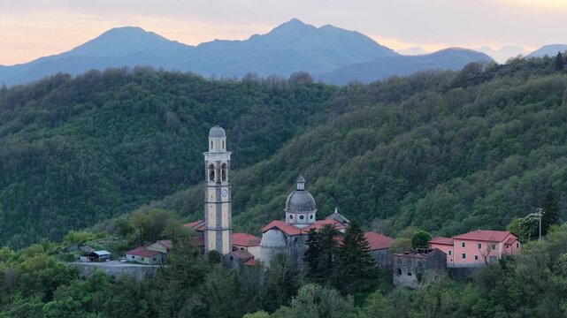 Aerial view of a historic church and bell tower nestled in the lush green mountains near Passo Di Cento Croci at dusk in Varese Ligure, Liguria, Italy.