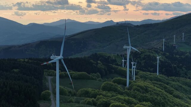 Aerial view of wind turbines on a mountain ridge at Passo di Cento Croci surrounded by lush green forests at sunset in Varese Ligure, Liguria, Italy.