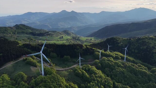 Aerial view of wind turbines at Passo Di Cento Croci on a green mountain ridge with rolling hills and forests under a soft sky in Varese Ligure, Liguria, Italy.