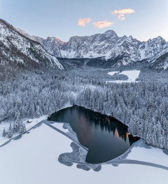 Winter landscape, aerial view of frozen lake and snow covered forest with Mangart mountains chain in the background, tourism destination concept, Fusine lakes, Tarvisio, Friuli, Italy.