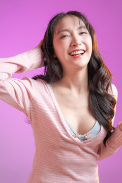 cheerful young East Asian woman with a vibrant surprised facial expression, holding her hands to her face and smiling joyfully. She is wearing a pink striped long-sleeve top against a solid pink studi