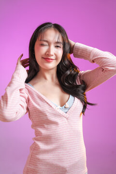 cheerful young East Asian woman with a vibrant surprised facial expression, holding her hands to her face and smiling joyfully. She is wearing a pink striped long-sleeve top against a solid pink studi