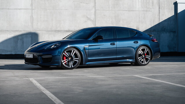 blue Porsche Panamera in front of concrete wall on sunny day. Three quarter low-angle front view of german grand tourer in rooftop parking lot. Kyiv, Ukraine - April 04 2026