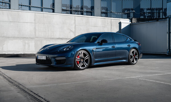 blue Porsche Panamera in front of concrete wall on sunny day. Three quarter low-angle front view of german grand tourer in rooftop parking lot. Kyiv, Ukraine - April 04 2026