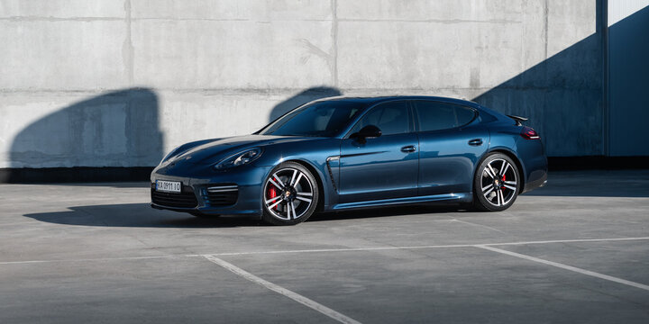 blue Porsche Panamera in front of concrete wall on sunny day. Three quarter low-angle front view of german grand tourer in rooftop parking lot. Kyiv, Ukraine - April 04 2026