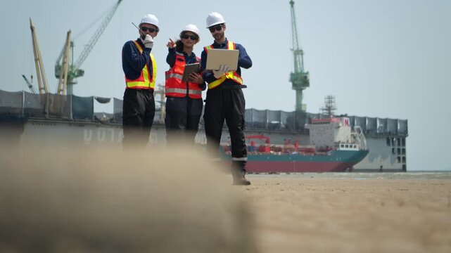 A team of engineers in safety gear walk beside the water at a port inspected the operations of the deep-sea port and cargo ships, Large ships and cranes are visible in the background