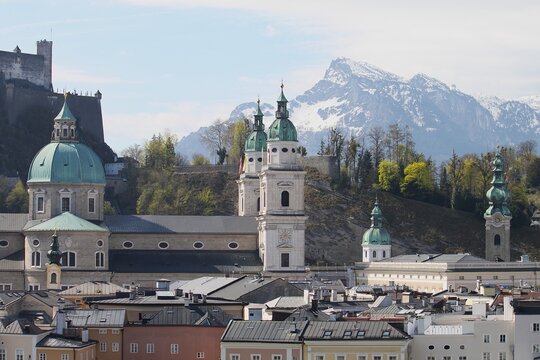 Salzburg Cathedral and Saint Peter Abbey domes with Hohensalzburg Fortress on Moenchsberg hill overlooking historic oldtown roofs and snow capped Untersberg mountain peak in Austria during sunny day.