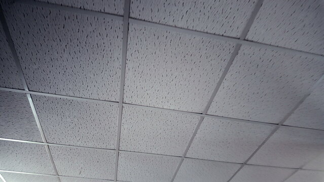 White acoustic drop ceiling with square perforated tiles in grid layout, viewed from below at diagonal angle inside commercial or institutional indoor setting