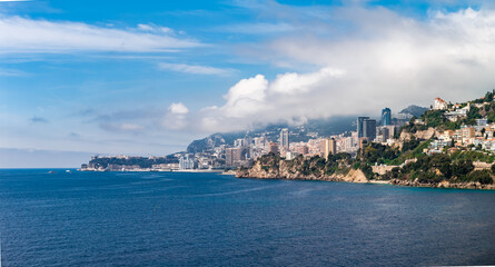 Fototapeta premium Monaco skyline hugs coastal cliffs on the French Riviera. Le Rocher sits left, Tour Odeon and beaches right, terraced villas and corniche roads frame under morning light.
