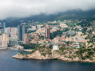 Fototapeta premium Aerial view of Monaco on the French Riviera shows towers and terraced apartments on steep hills, a curved bay, pale cliffs, a white villa, and turquoise water under overcast skies.