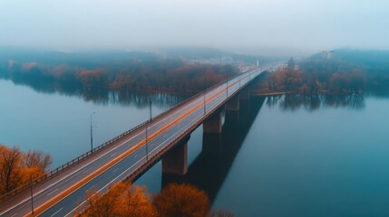 Obraz premium A foggy bridge stretches across a calm river, lined with autumn-colored trees along the banks