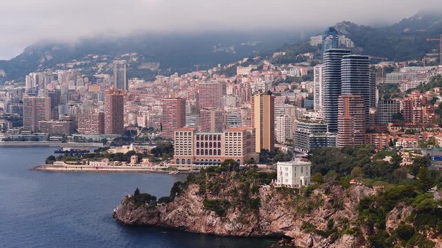 Aerial view of Monte Carlo, Monaco, with high rise towers, a seaside hotel, a white villa on a rocky point, cranes, boats, and haze as the camera sweeps slowly.