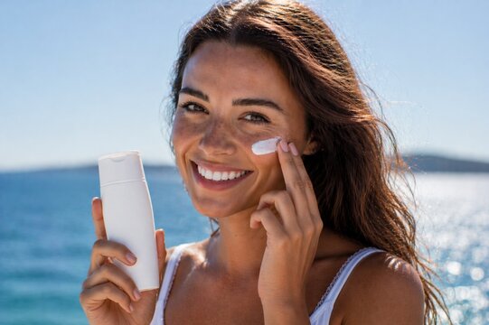 Beautiful young woman applying sunscreen cream on her cheek while holding a blank cosmetic bottle. Shot at the beach with bright sunlight and natural skin texture. Perfect for skincare, UV protection,