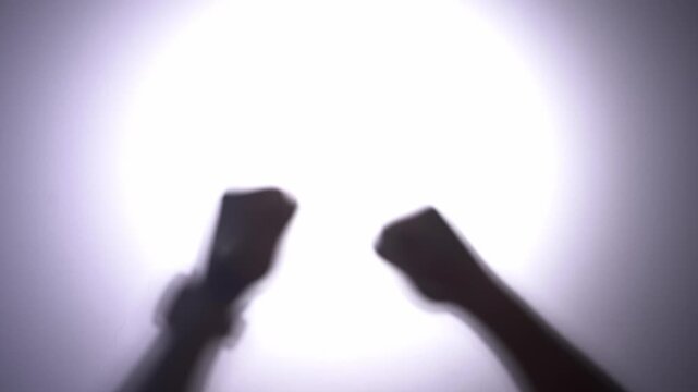Bright circular light dominates the background. Two silhouetted figures emerge from it. One holds a smartphone. Another raises a fist. Their gestures suggest triumph and excitement
