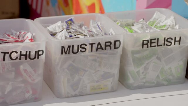 This close up, panning video shows ketchup, mustard, and relish condiments in containers at an outdoor vendor's stand. 