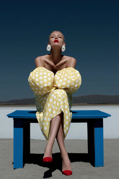 A female model in yellow polka dot dress sits on blue table in desert landscape. Elegant retro fashion concept with dramatic styling.
