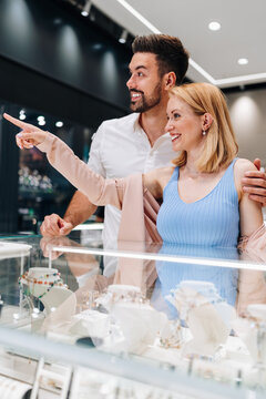 A happy young couple looks at jewelry in a glass display case at a luxury retail store. The woman points at a piece of jewelry while the man smiles and embraces her.