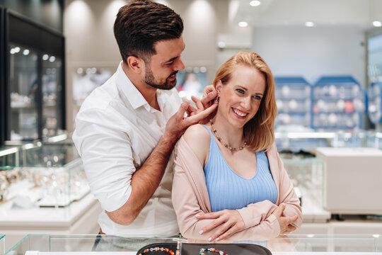 A man helps his smiling partner try on elegant earrings while shopping together in a modern jewelry store with glass display cases.