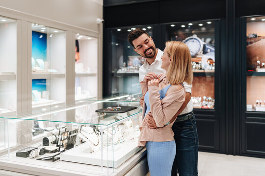 A happy couple chooses a diamond necklace in a high-end jewelry boutique. The man embraces the woman as they admire fine jewelry in a glass display case.