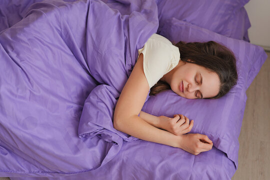 A young woman is lying on her side in a bed. She is covered with a purple blanket and appears to be sleeping. Natural light fills the room