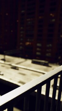 A person stands on a balcony overlooking the city skyline at night. The lights of nearby buildings shine, creating a busy urban atmosphere. The cold air invites moments of reflection.