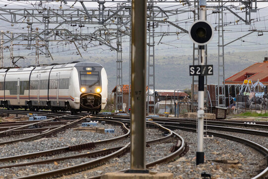 Commuters and travellers gather along the platforms at &Aacute;vila railway station as a regional train arrives, framed by overhead cables and urban architecture