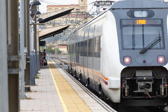 Commuters and travellers gather along the platforms at &Aacute;vila railway station as a regional train arrives, framed by overhead cables and urban architecture