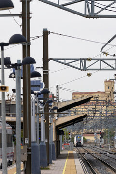 Commuters and travellers gather along the platforms at &Aacute;vila railway station as a regional train arrives, framed by overhead cables and urban architecture