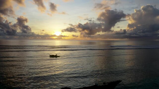 Aerial view of a lone fisherman in a traditional outrigger boat on the calm ocean surface during a golden sunset with dramatic clouds in Morotai, Indonesia.