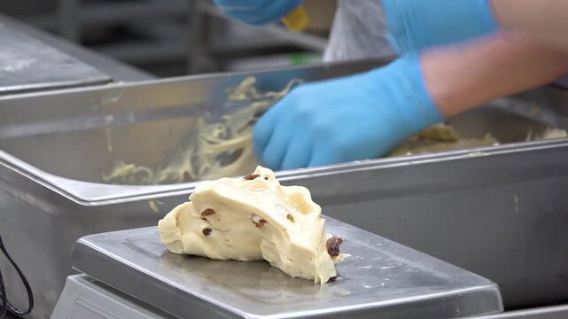Detailed footage of the dough scaling process in a food factory. A worker separates pieces of yellow dough filled with raisins and places them on a digital scale, demonstrating quality control and sta