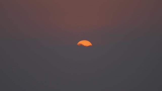 Timelape orange sun hangs low in a hazy sky, surrounded by warm amber tones and soft mist, creating a calm minimalist sunset scene with muted gradients and tranquil mood.