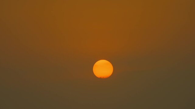 Timelape orange sun hangs low in a hazy sky, surrounded by warm amber tones and soft mist, creating a calm minimalist sunset scene with muted gradients and tranquil mood.