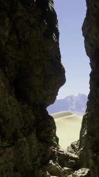 A gap between two large rock formations shows a shape that resembles a human face. In the background, mountains rise and sand dunes stretch under a clear sky.