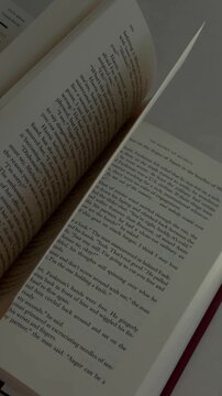 Handheld slow motion tracking pan of hand turning pages of paperback book on table in indoor study during daytime, quiet reading scene