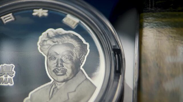 Cinematic close up video of a Fukushima Prefecture 1000 yen silver coin featuring the portrait of Hideyo Noguchi and prominent Fukushima lettering, presented inside a protective capsule.