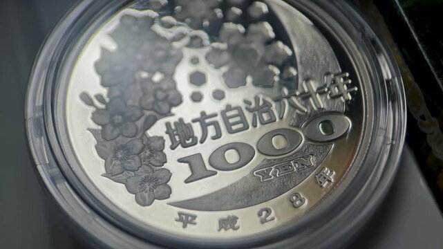 Close up video of the reverse side of a Japanese 1000 yen silver coin featuring floral cherry blossom elements, bold denomination, and Japanese lettering.