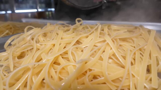 Close-up shot of a chef draining hot, freshly boiled linguine pasta in a stainless steel colander over a metal baking sheet with some water and oil. The steam is rising from the hot pasta. This footag