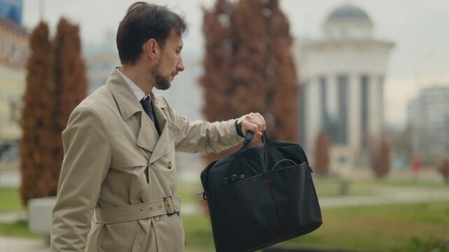 A businessman checks documents and his watch while waiting for an outdoor business meeting. The professional prepares for a rendezvous in an urban park setting.