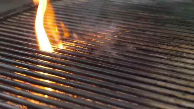 A close-up shot of a red, bristle-free grill brush being used to clean a hot, flaming barbecue grill grate. The brush effectively removes residue from the stainless steel grates, ensuring a clean and 