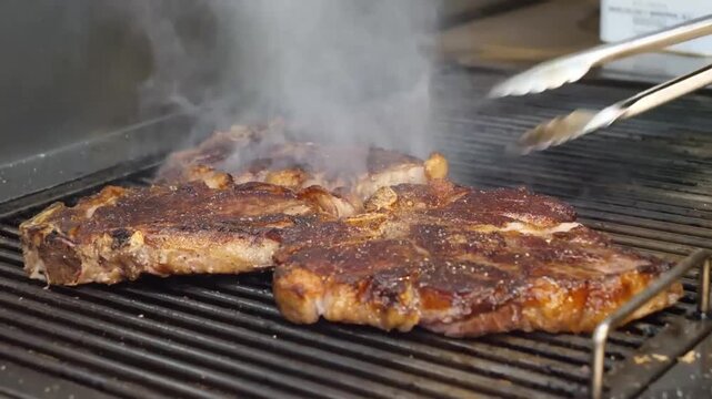Two seasoned T-bone steaks are seared to perfection on a metal outdoor grill. Plumes of aromatic white smoke rise from the juicy meat as it cooks over high heat, showcasing beautiful grill marks and c