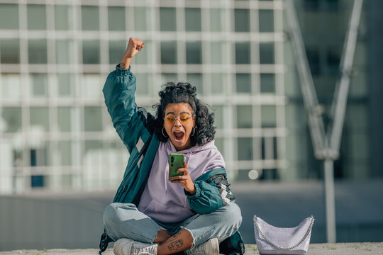 A euphoric young woman with curly hair raises her fist in a victory gesture while checking her phone, sitting cross-legged in front of a modern glass building: energy, freedom, and digital triumph