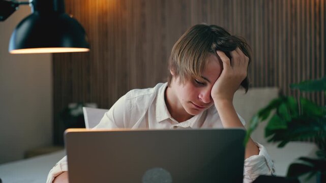 Overwhelmed and exhausted teenage boy struggling with homework, falling asleep at his desk late at night while using laptop for e-learning, feeling tired and rubbing his eyes.