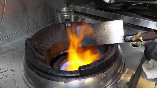A dynamic close-up of a high-power commercial gas burner igniting a large orange and blue flame, demonstrating the "wok hei" technique in a professional restaurant kitchen.