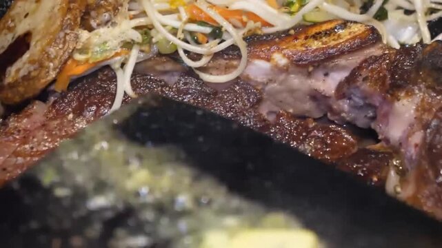 A close-up shot of a pat of herb butter sizzling and melting on a hot black griddle or pan, next to a piece of cooking steak. The vibrant yellow butter bubbles as it liquefies, ready for basting or co