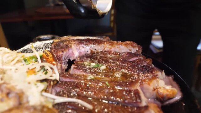 A high-definition, close-up shot of a large, perfectly cooked ribeye steak sizzling on a cast-iron griddle in a restaurant setting. A metal spatula or cleaver is used to slice the tender, juicy meat. 