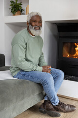 Naklejka premium Senior African American man sitting on sofa in living room wearing sweatshirt smiling by fireplace