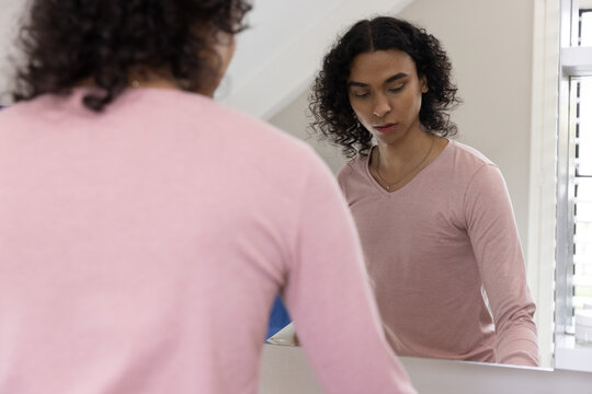 Non-binary person in pink V-neck and chain, standing and looking into vanity mirror with blue towel