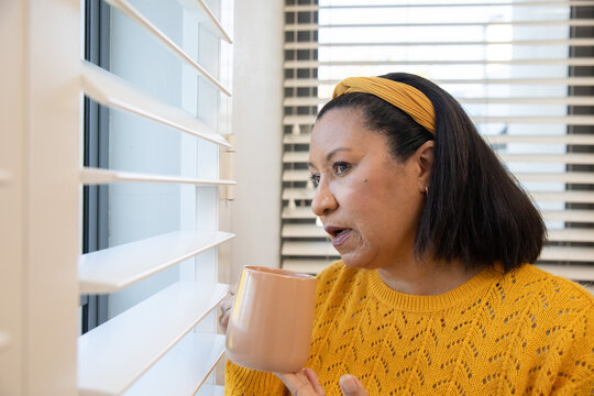 Mature woman sitting by window in yellow sweater and headband with peach mug, gazing through blinds