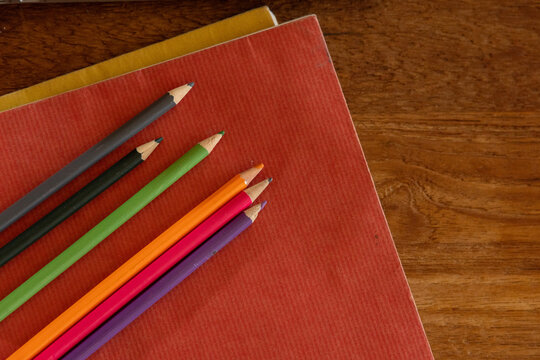 Seven pencils resting diagonally on red notebook with yellow edge on left wooden desk, copy space