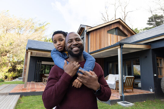 African American dad carrying son piggyback on lawn in front of house, maroon sweater, denim jacket
